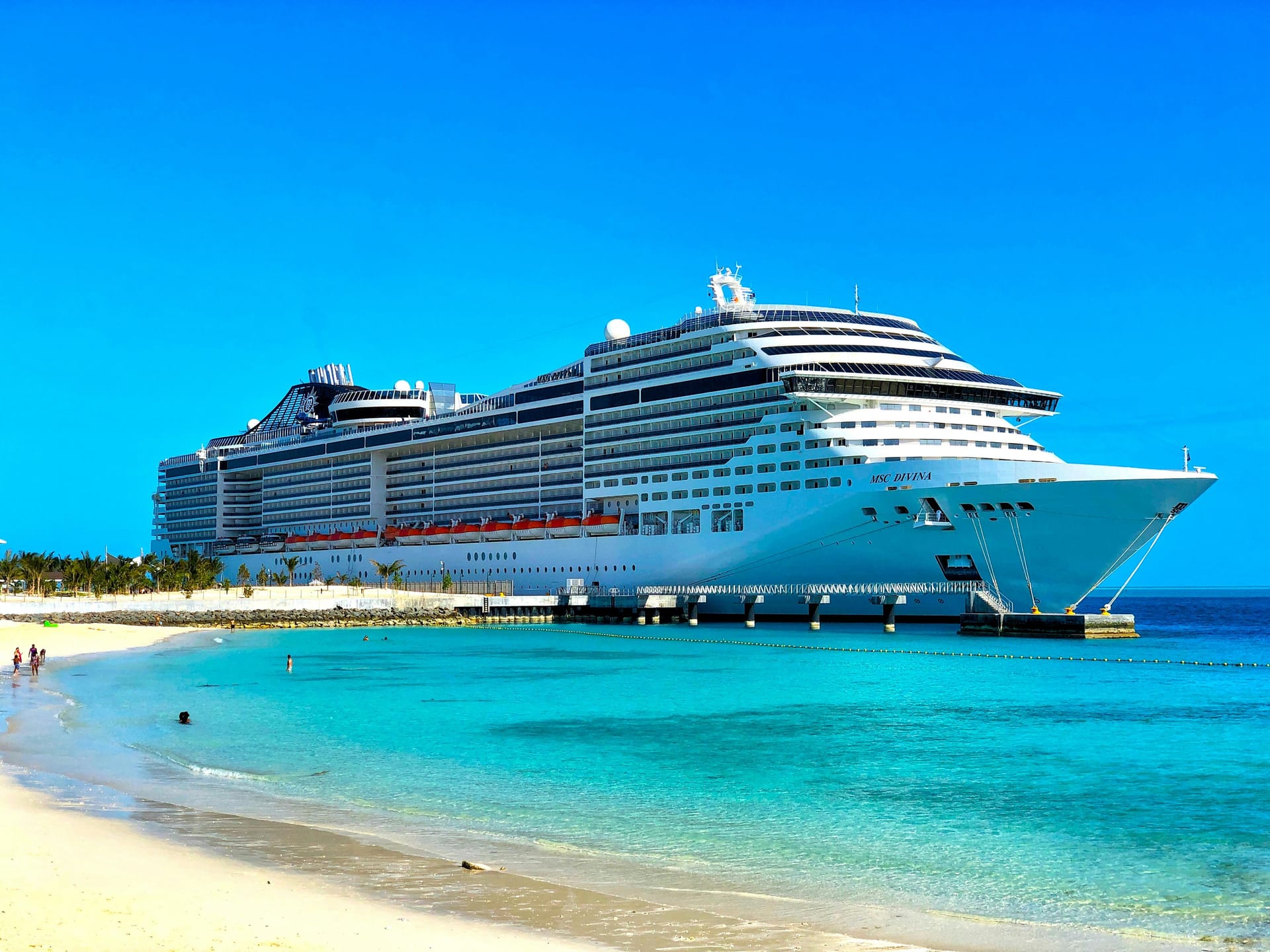 A massive white cruise ship docked at a tropical island with turquoise water and a sandy beach in the foreground.