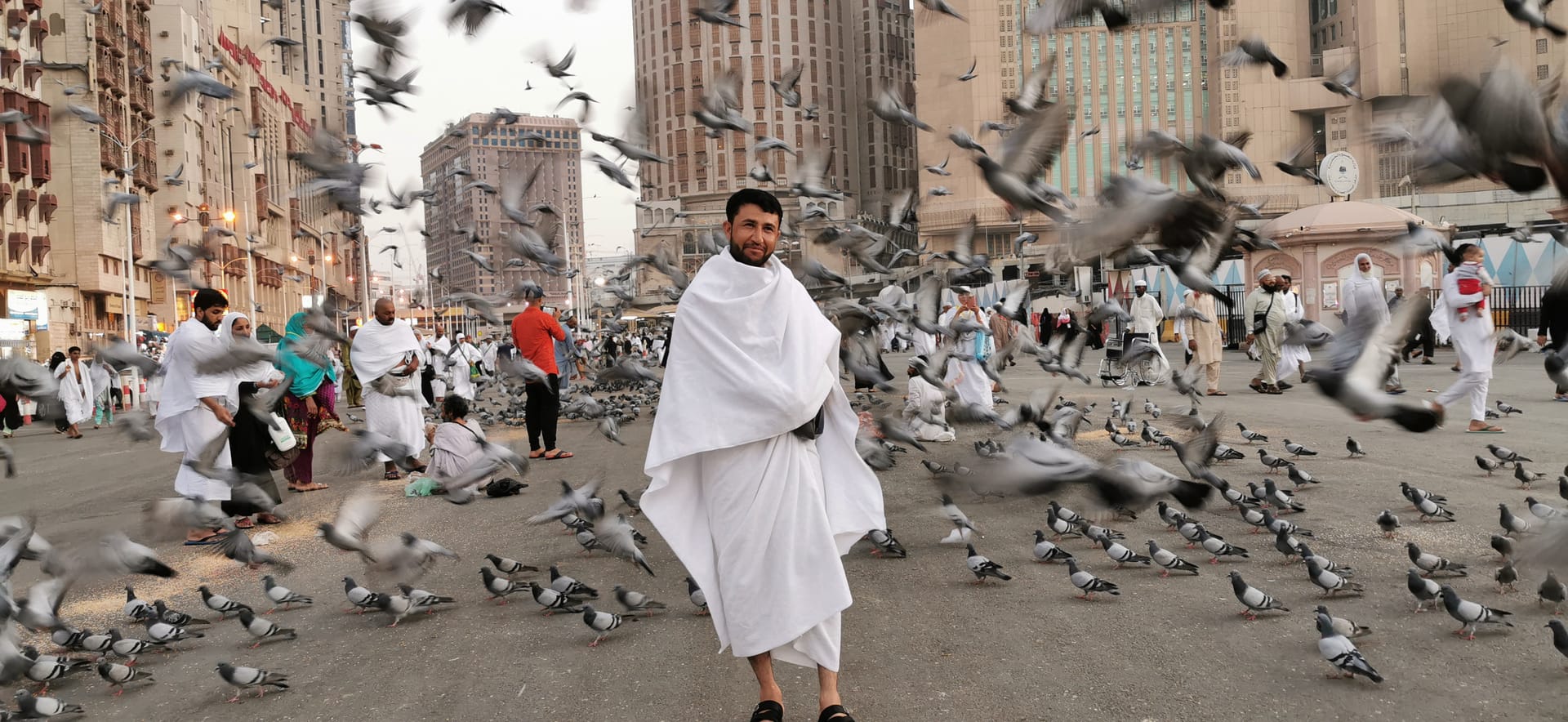A pilgrim wearing white Ihram robes walks through a large flock of pigeons on a city street.