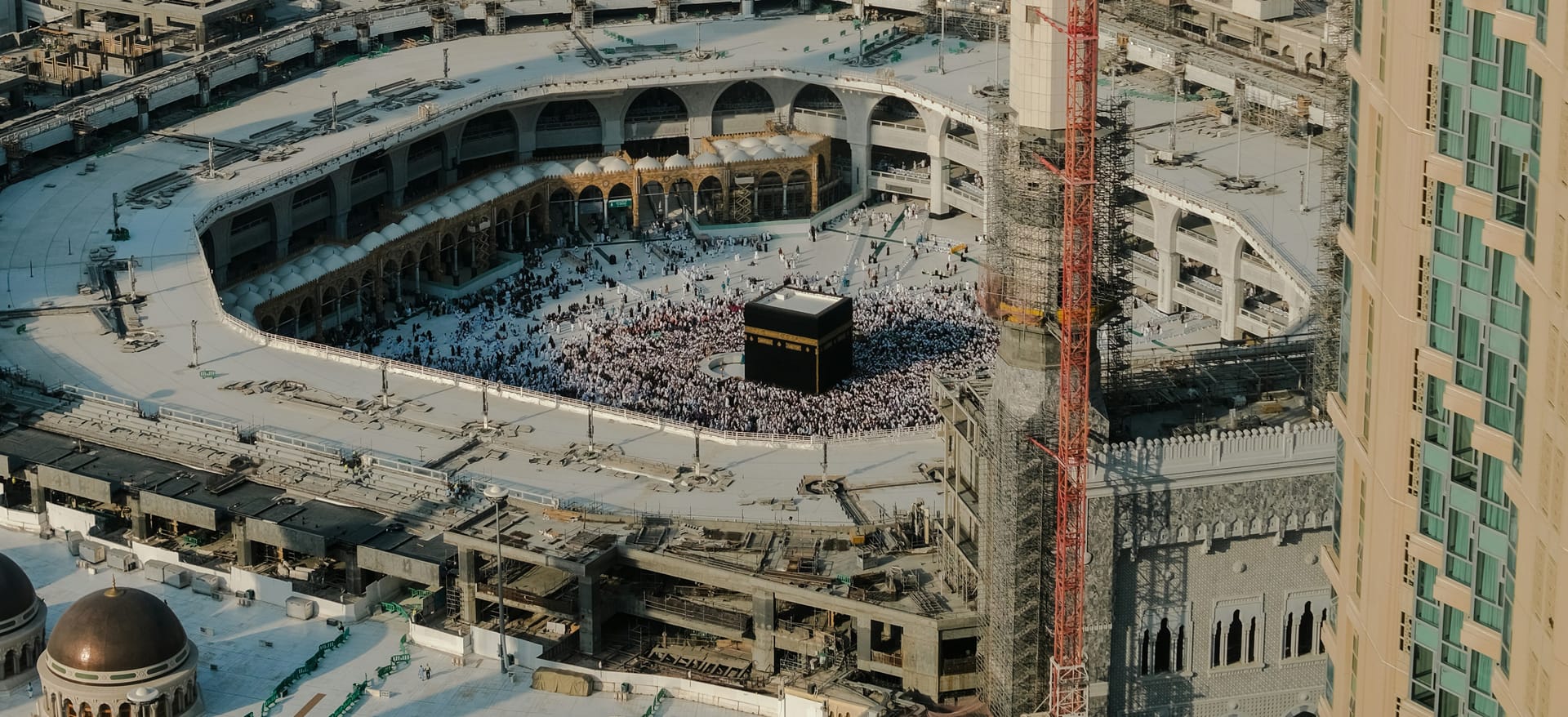 An aerial perspective of the Grand Mosque in Mecca showing the Kaaba, the courtyard, and the surrounding multi-story architecture and minarets.
