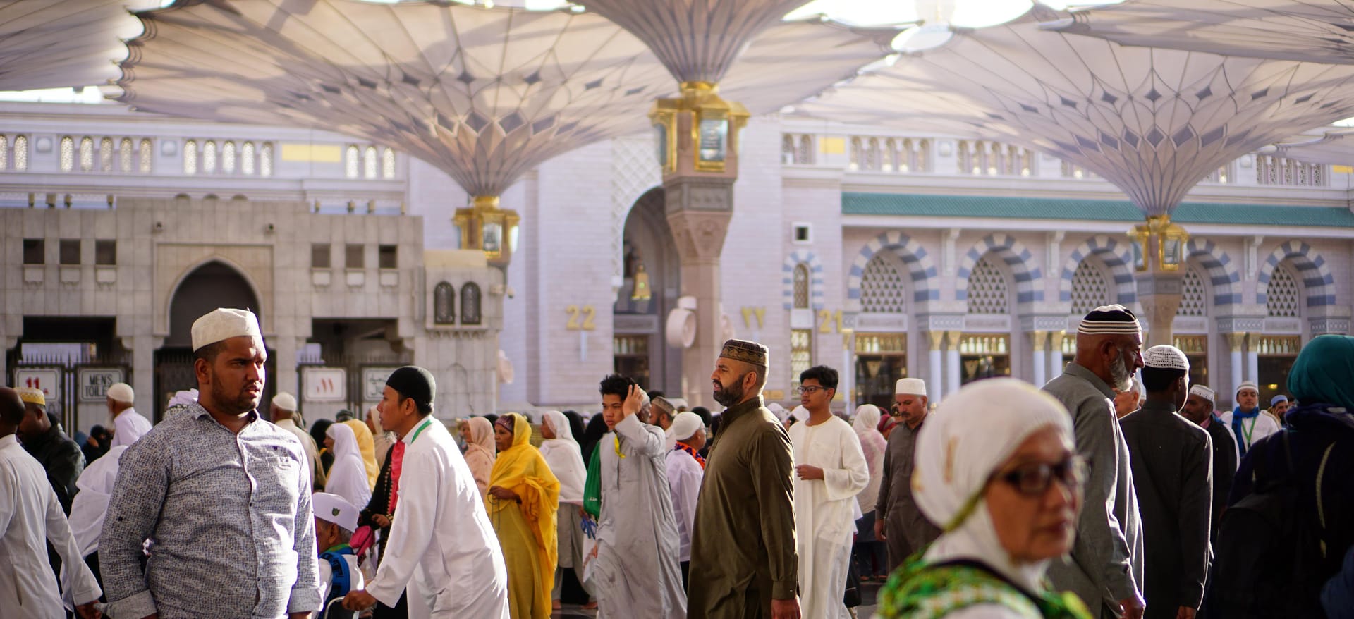 A diverse crowd of pilgrims walking through the courtyard of the Prophet's Mosque in Medina, with large umbrellas visible in the background.