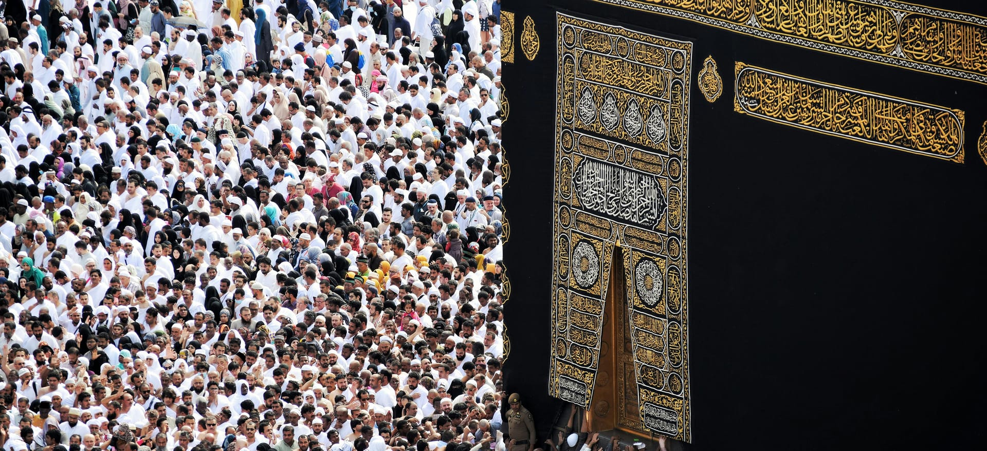 High-angle close-up view of a dense crowd of pilgrims pressing against the black and gold cloth of the Kaaba to touch it.