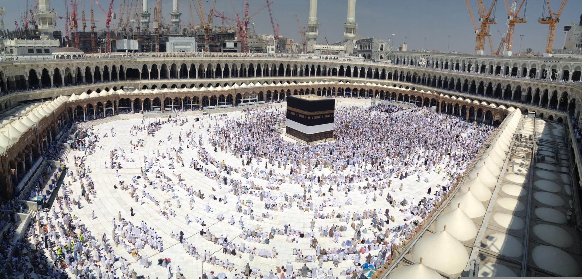 A wide aerial shot of the white marble courtyard of the Great Mosque of Mecca filled with worshippers gathered around the Kaaba.