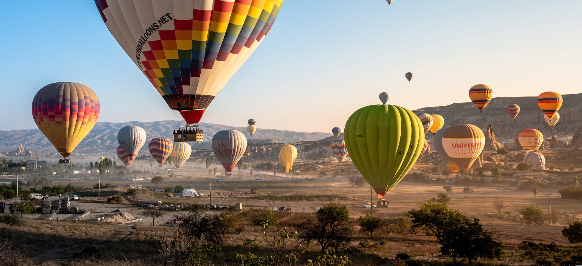 Dozens of colorful hot air balloons flying over the arid, rocky landscape of Cappadocia at sunrise.