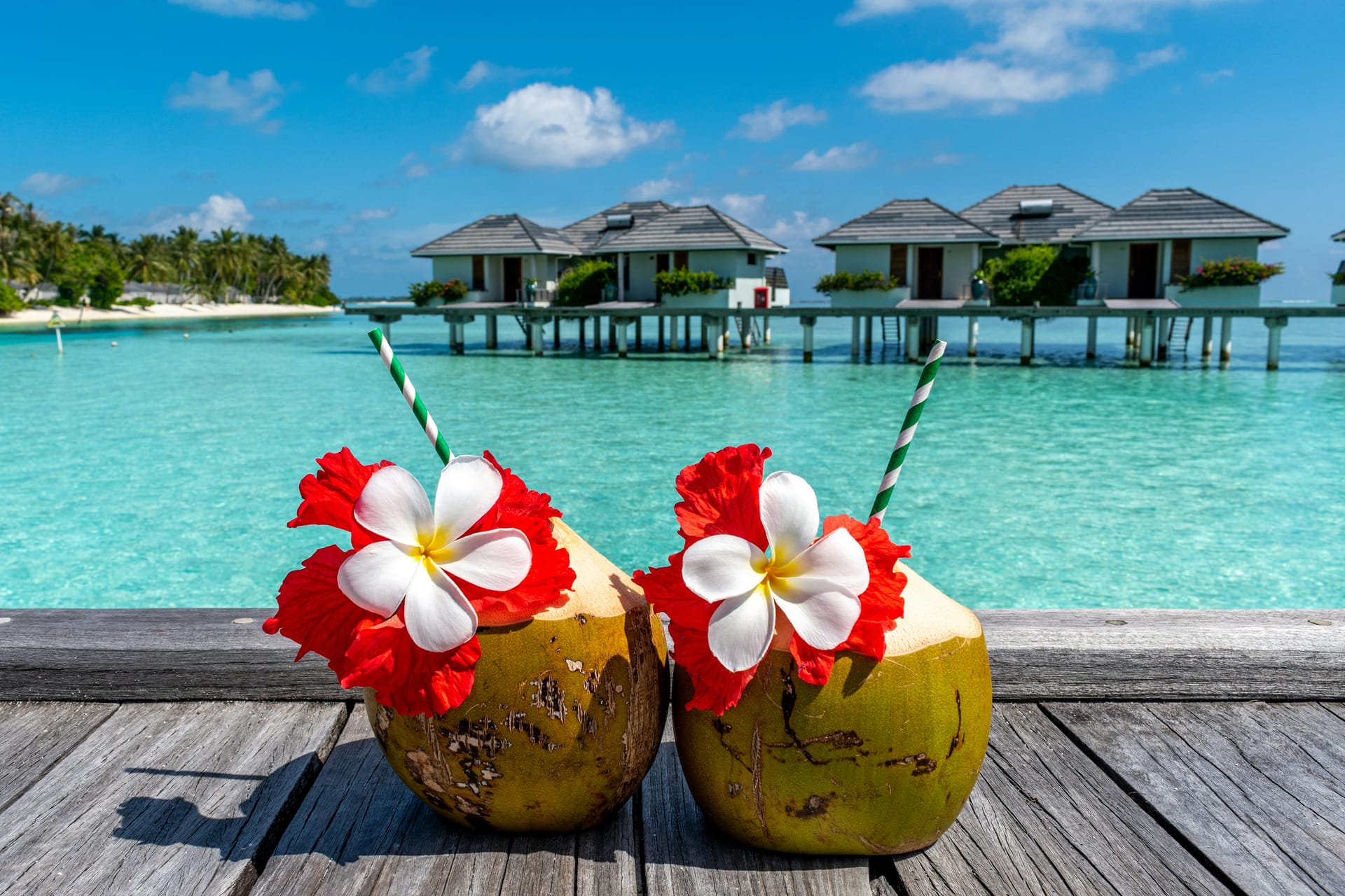 Two fresh coconuts decorated with red and white flowers sitting on a wooden deck, with overwater bungalows and blue ocean in the background.