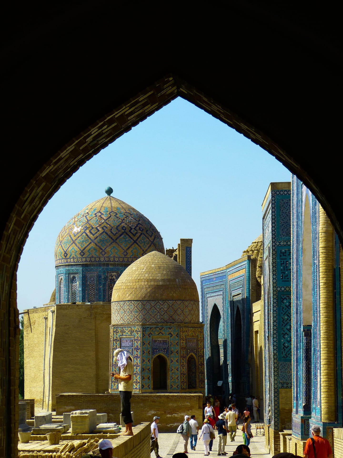 A view through a dark stone archway revealing the bright blue tiled domes and mausoleums of the Shah-i-Zinda necropolis under a clear sky.