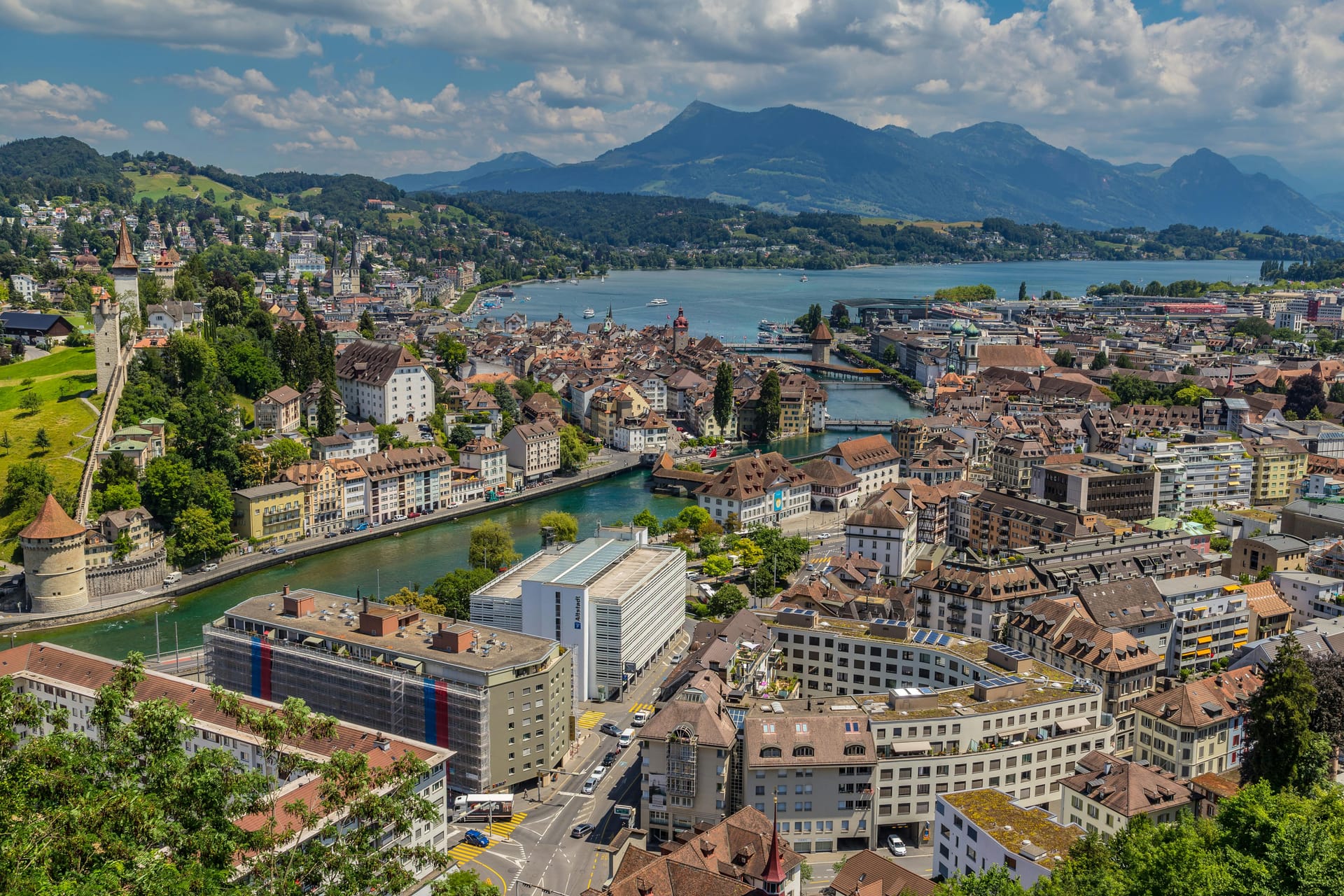 A panoramic cityscape of Lucerne, Switzerland, showing the Reuss River flowing through the town and the historic city walls on the green hillside.