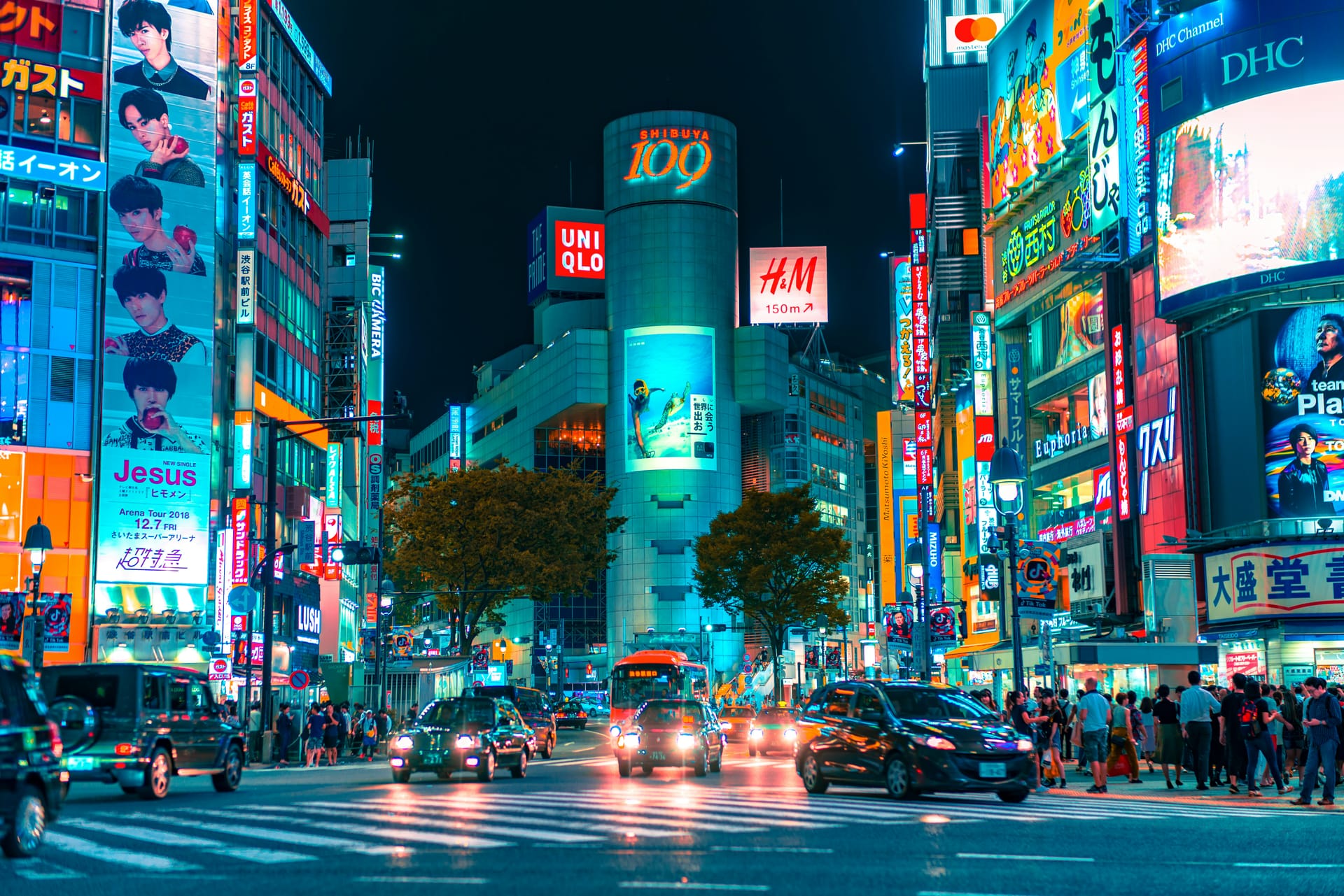 A busy street scene in Shibuya, Tokyo at night, filled with bright neon signs, advertisements, and cars.