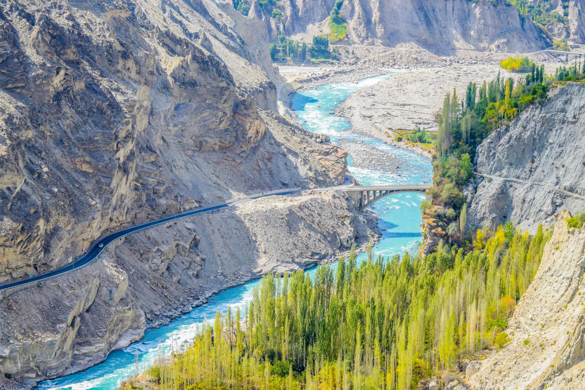A turquoise river winding through a deep rocky gorge in the Hunza Valley, with a road carved into the cliffside and green trees along the bank.