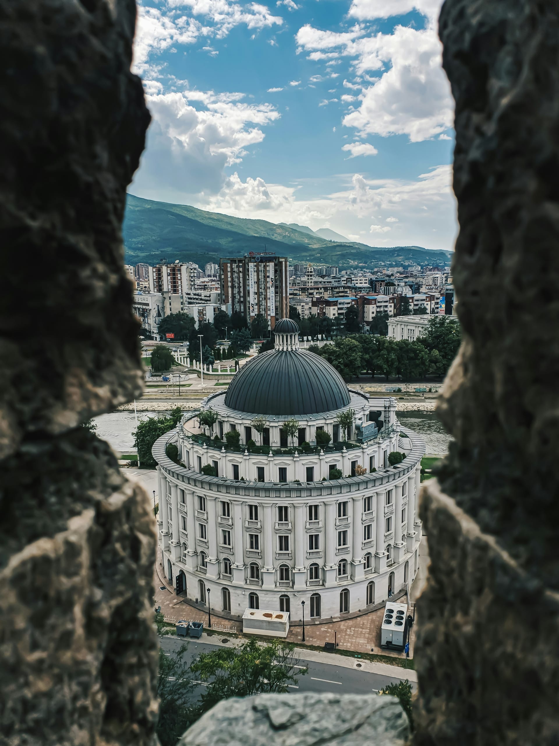 A view of a white classical domed building in Skopje framed by a dark stone window or archway.