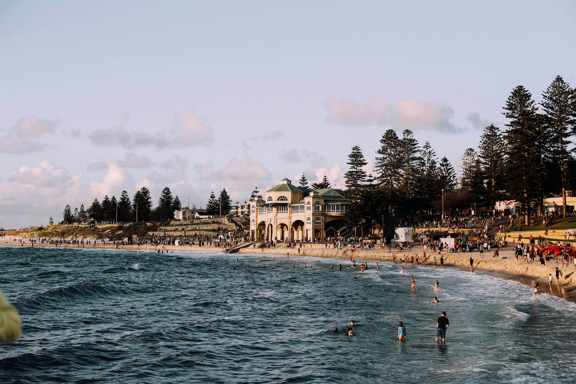 People swimming and relaxing on the sand at Cottesloe Beach with the historic Indiana Teahouse building in the background.