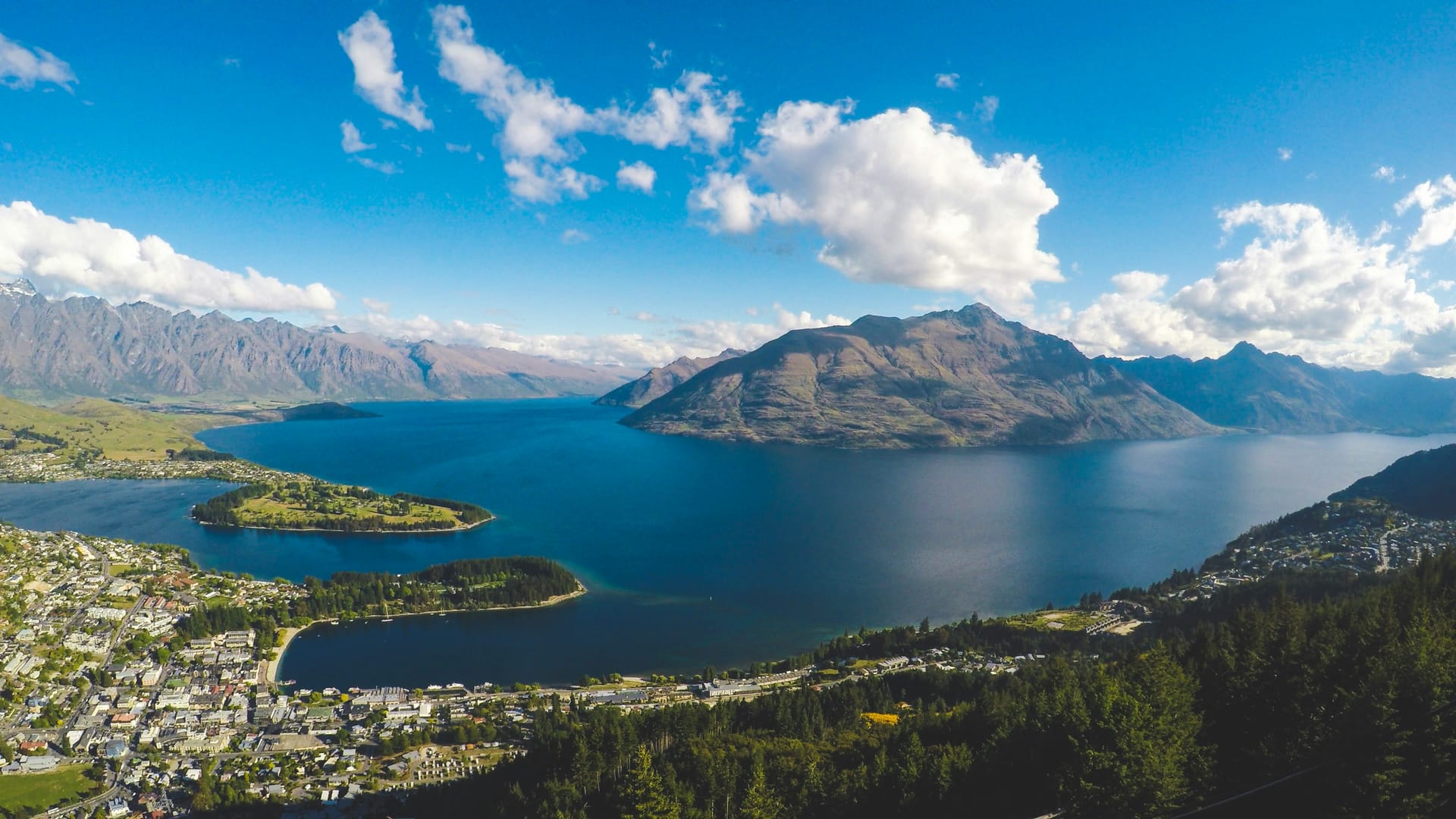 A high-angle panoramic view of a town nestled on a peninsula jutting into a large blue lake, surrounded by jagged mountains under a blue sky.