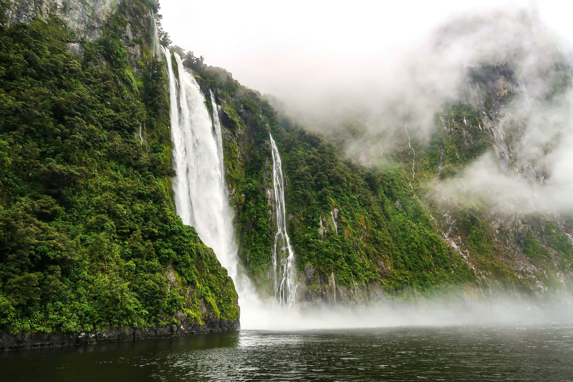 A powerful waterfall cascading down a steep, mossy cliff face into the dark water of a fjord, shrouded in mist.