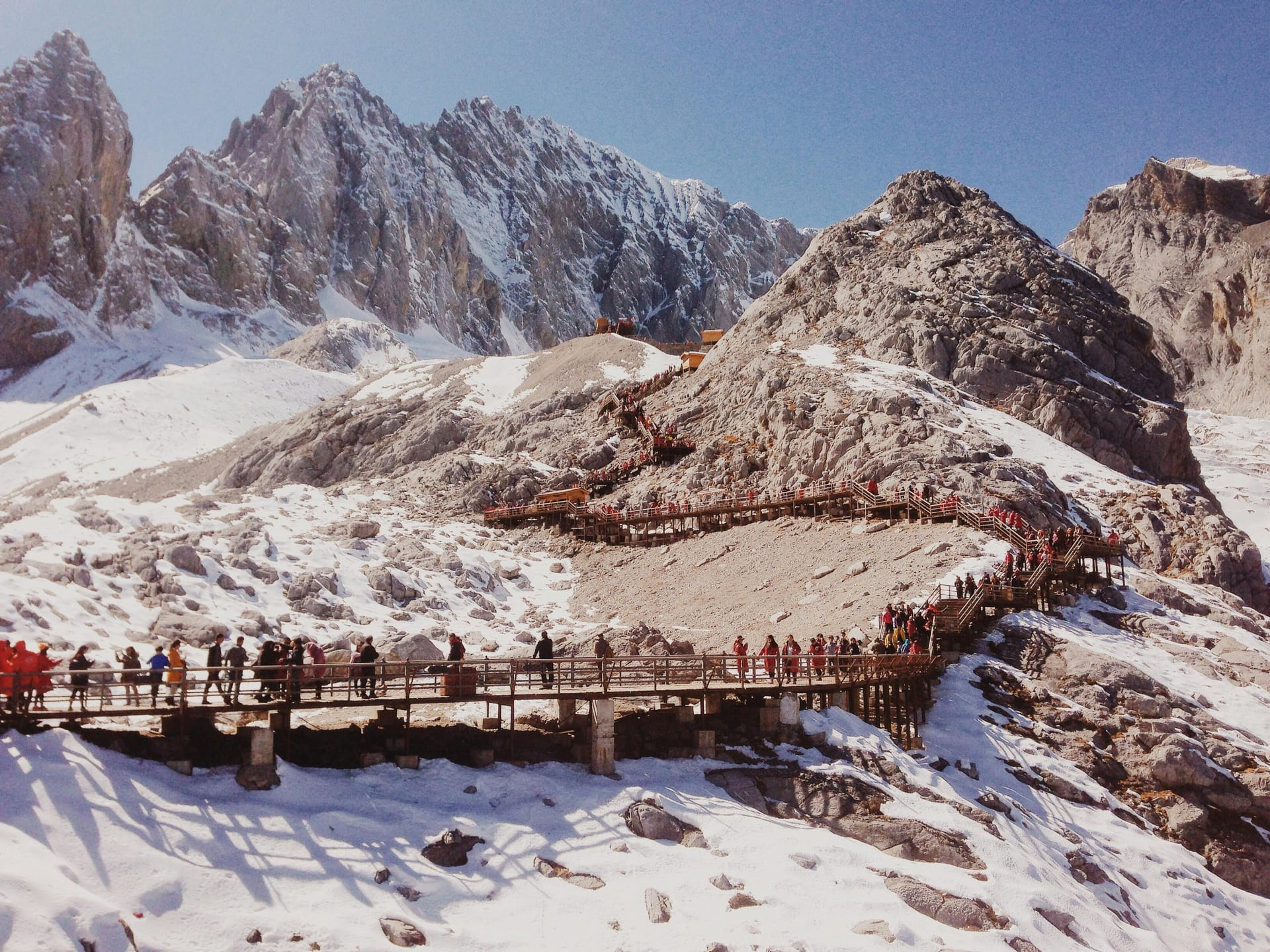 A wooden walkway crowded with tourists winding through the snowy landscape of Jade Dragon Snow Mountain with jagged peaks in the background.