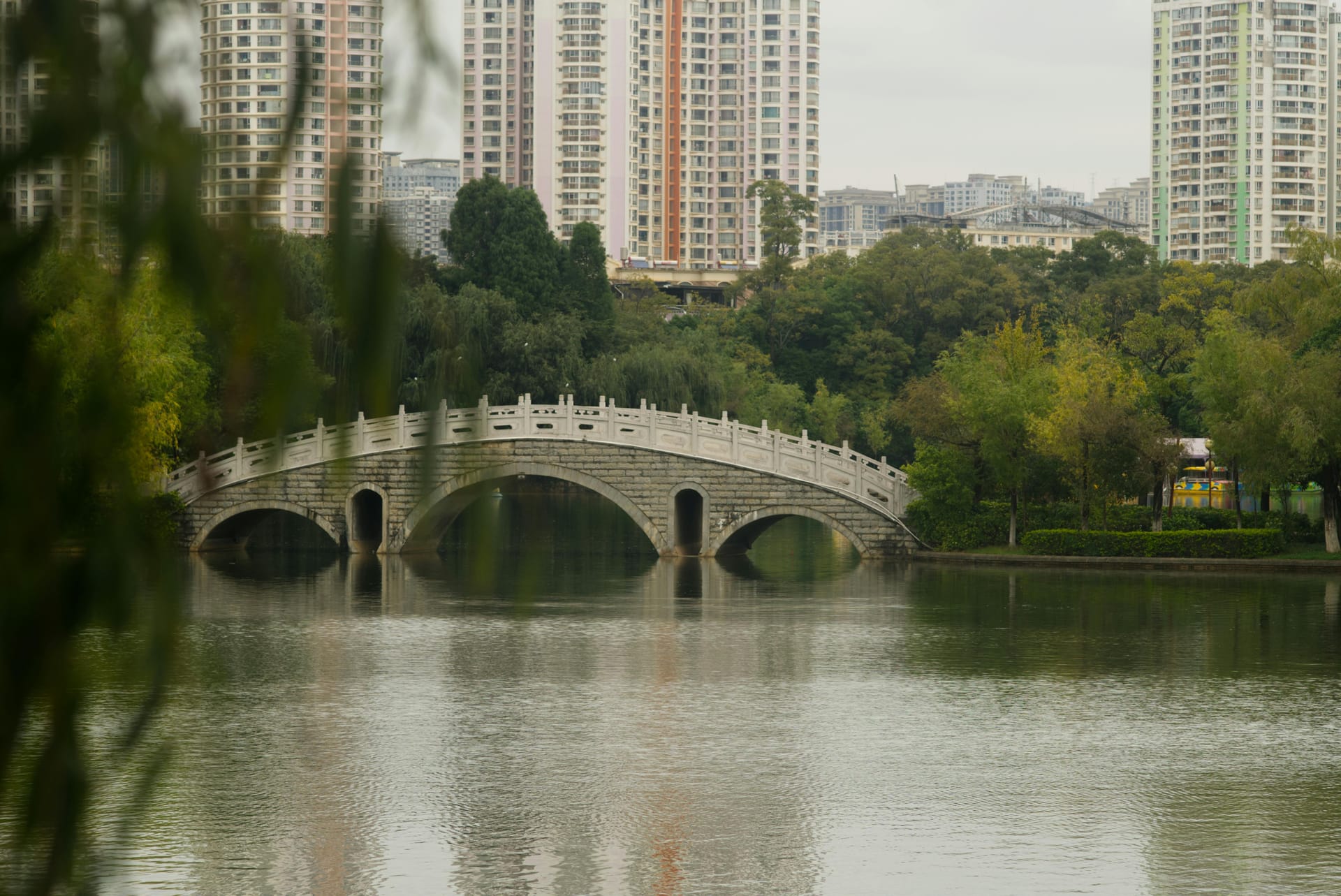 A stone arch bridge crossing the calm waters of Green Lake Park in Kunming, framed by weeping willow trees.
