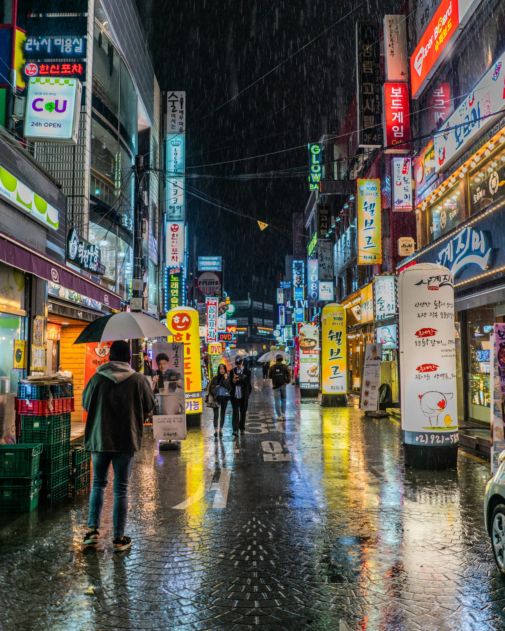 A rainy night street scene in a Korean city district filled with bright neon signs and pedestrians holding umbrellas.
