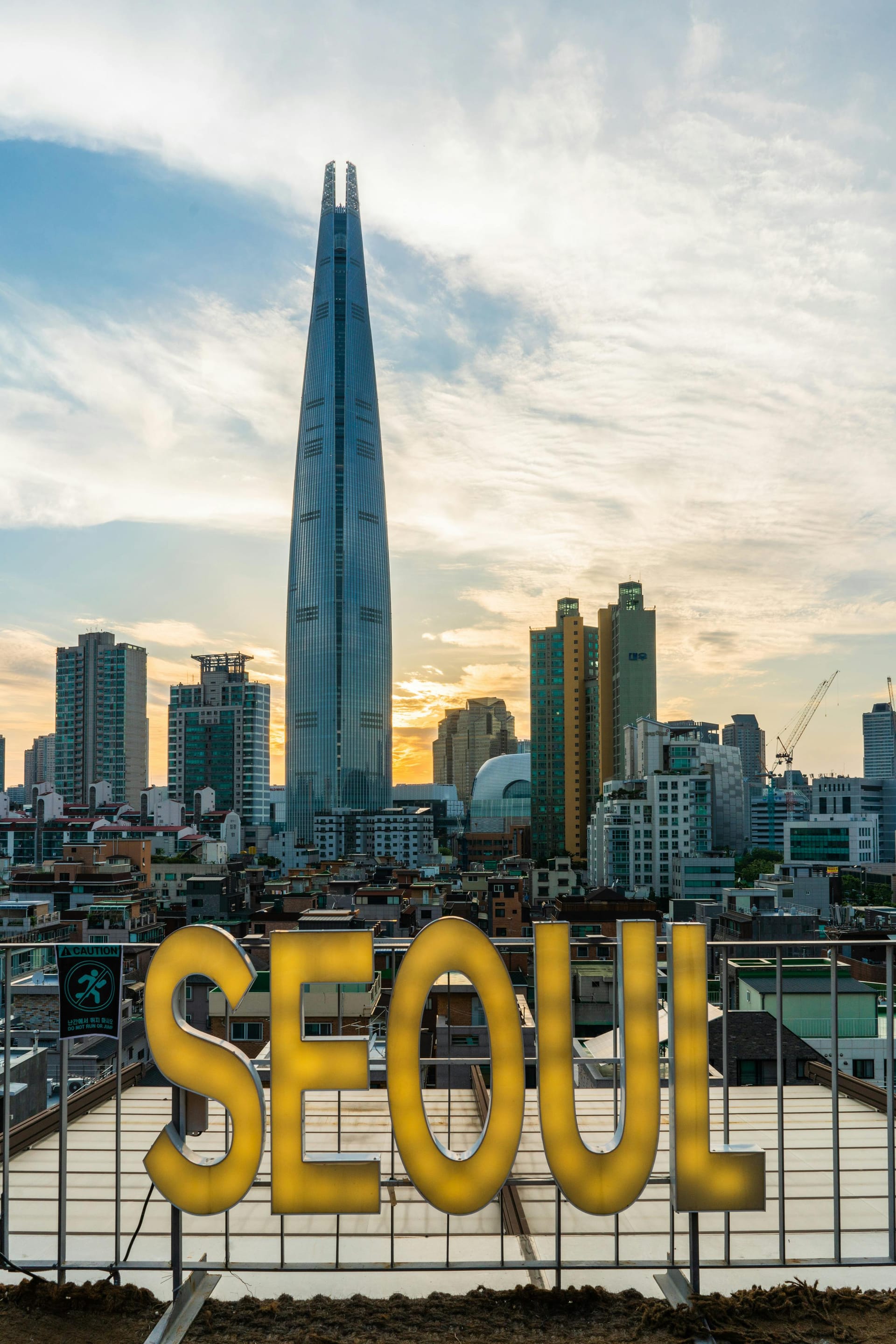 The towering Lotte World Tower skyscraper rising above the Seoul city skyline with large gold "SEOUL" text at the bottom.
