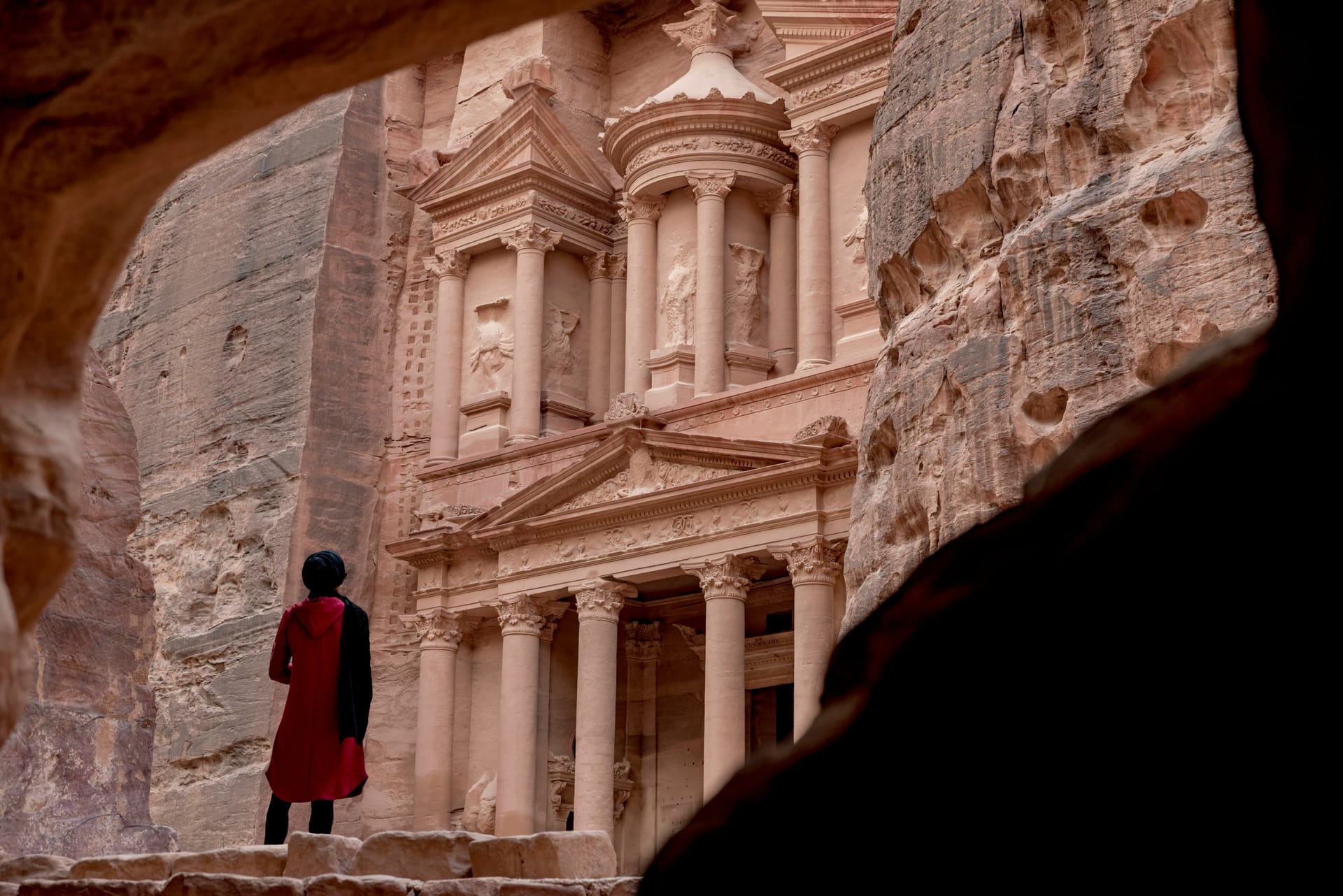 A person wearing a red shawl stands in front of the massive rock-cut facade of the Treasury (Al-Khazneh) in Petra.
