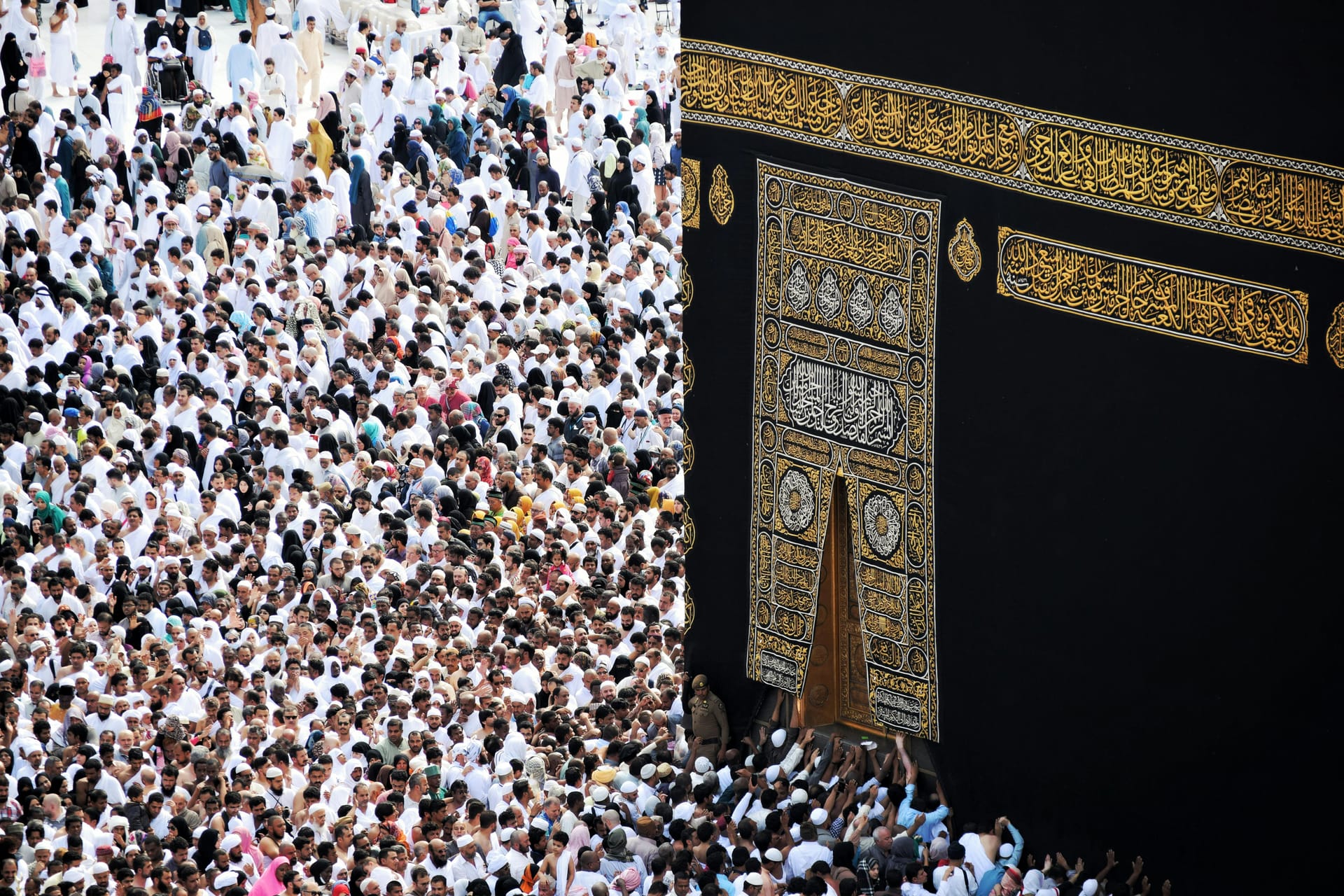 High-angle view of a dense crowd of pilgrims pressing against the black cloth and gold door of the Kaaba.