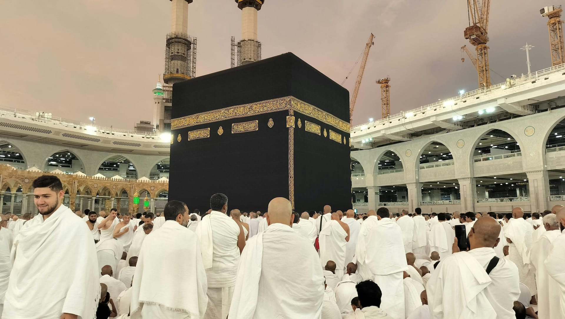 Male pilgrims wearing white Ihram robes gathering near the Kaaba in the illuminated courtyard of the Grand Mosque.