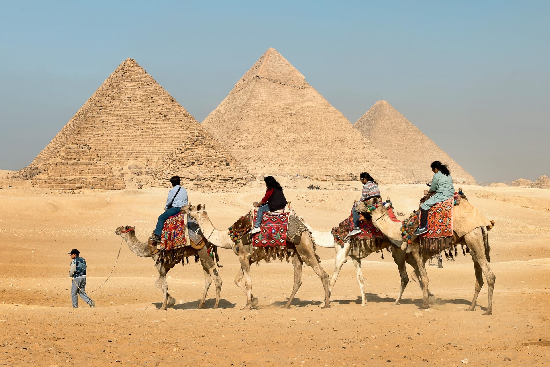 Tourists riding camels across the sand with the three Great Pyramids of Giza in the background.