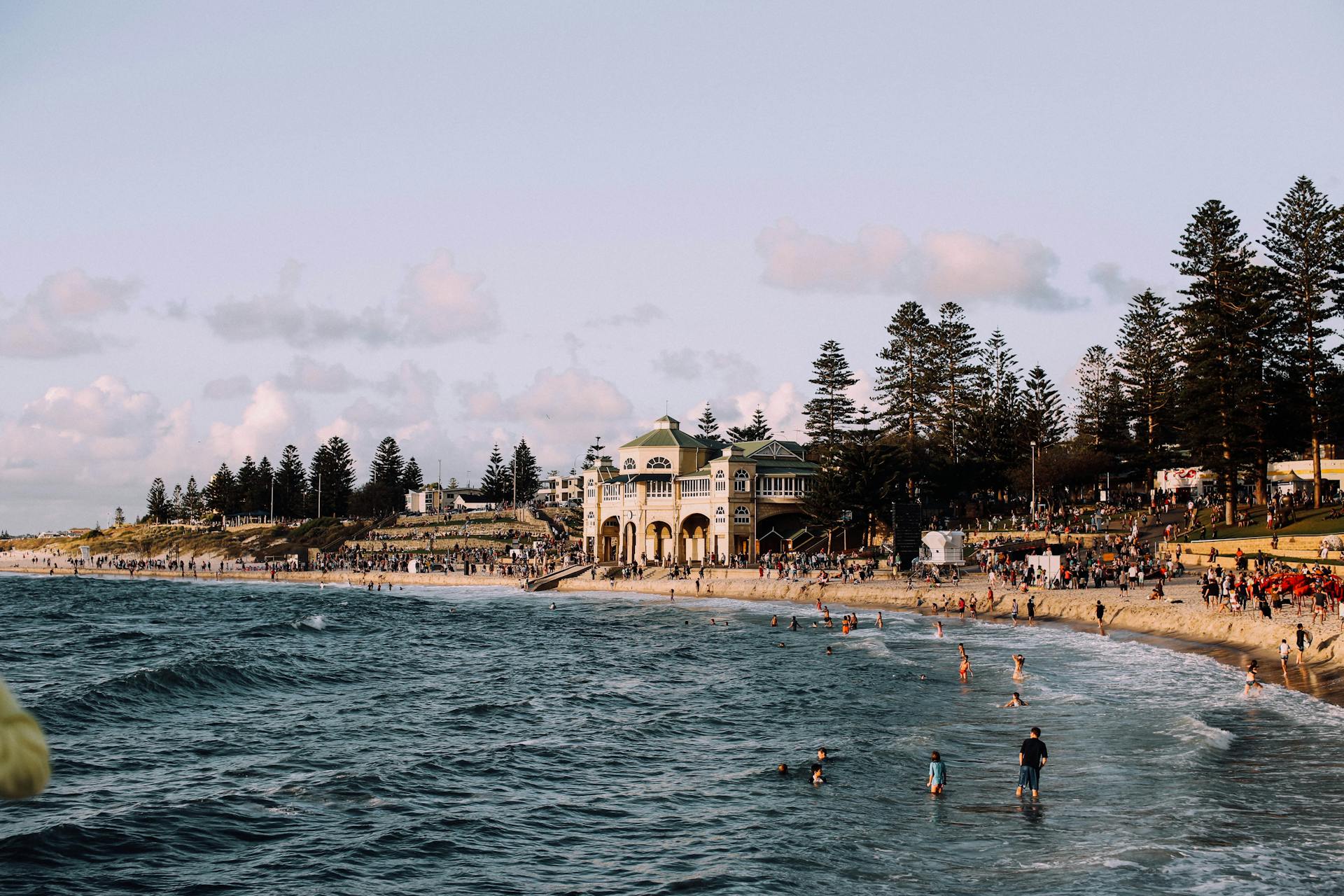 People relaxing on the sand and swimming in the ocean at Cottesloe Beach, with the historic Indiana Teahouse building and Norfolk pine trees in the background