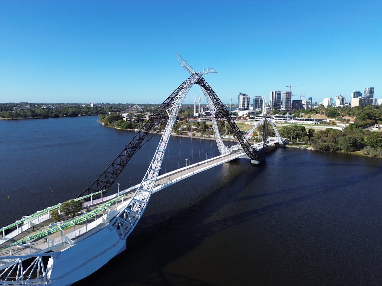 Aerial view of the white, curved arches of the Matagarup Bridge spanning the river, with the city skyline visible in the distance.