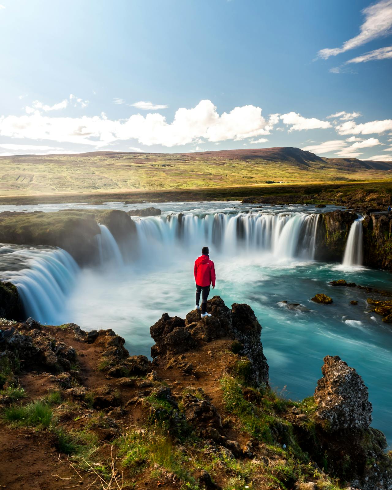 A person in a red jacket standing on a rocky outcrop looking at the wide, horseshoe-shaped Goðafoss waterfall under a bright blue sky.