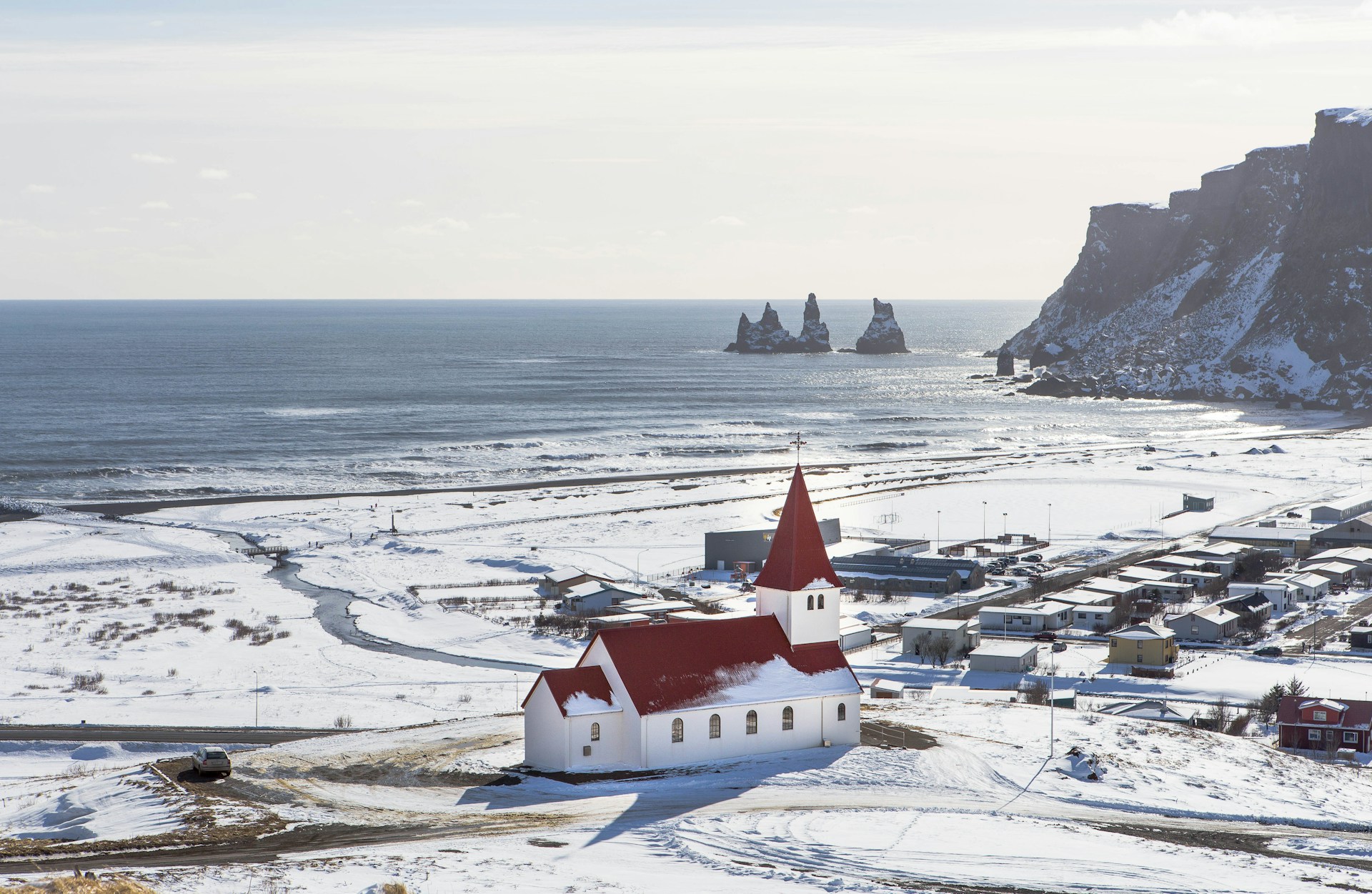 The red-roofed Víkurkirkja church stands out against a snowy landscape in the village of Vík, with the Atlantic Ocean and sea stacks in the distance.