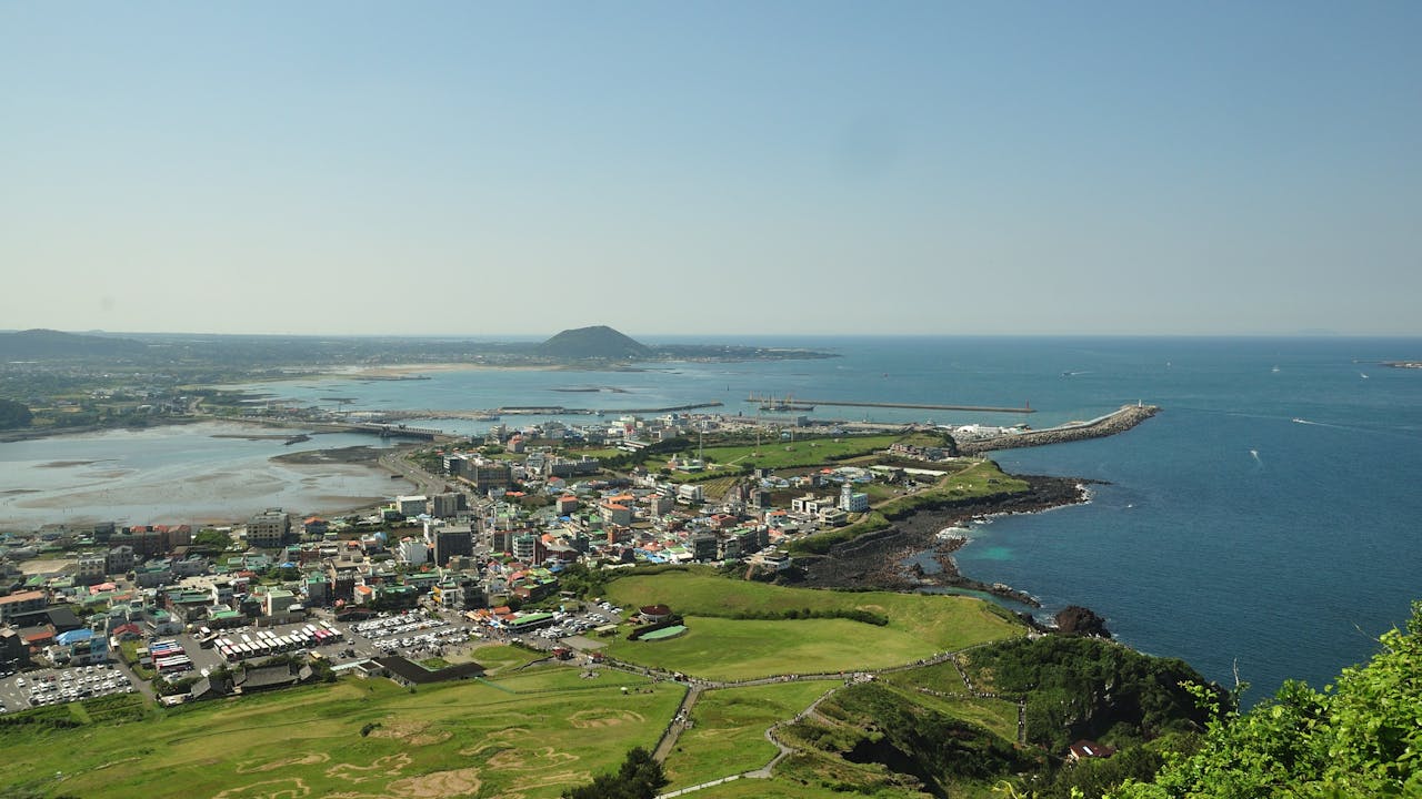 A panoramic view of the green coastline of Jeju Island with a volcanic hill rising in the distance under a blue sky.