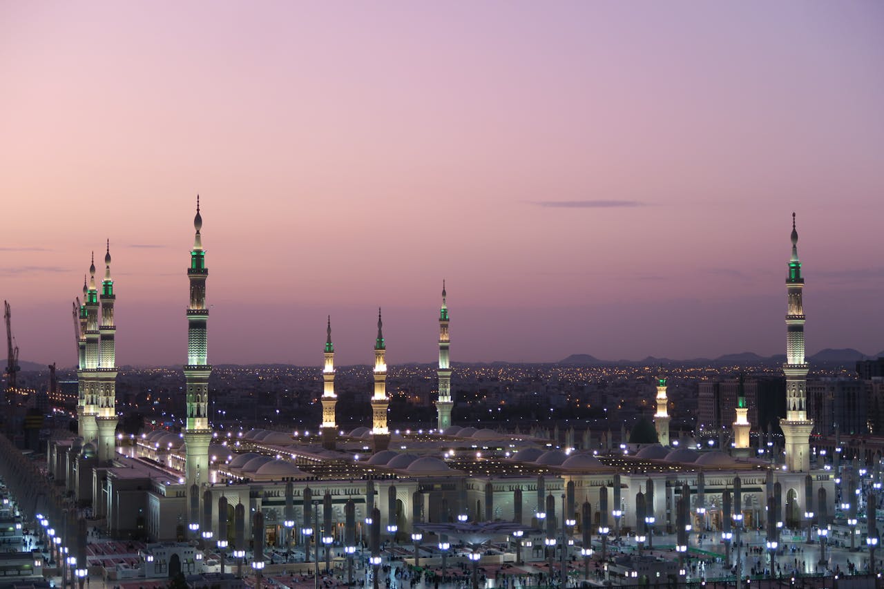 A wide panoramic view of the many illuminated minarets of the Prophet's Mosque in Medina against a purple twilight sky.